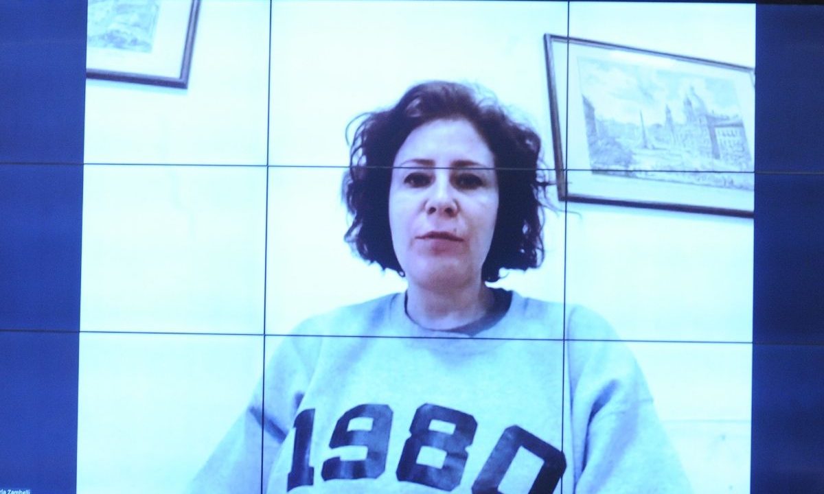 Foto de uma tela de monitor exibindo a ex-deputada federal Carla Zambelli em uma transmissão de vídeo. Ela está com cabelo curto e cacheado e veste um moletom cinza com '1980' estampado. Atrás dela, há dois quadros emoldurados com paisagens de Roma. A tela exibe linhas de grade