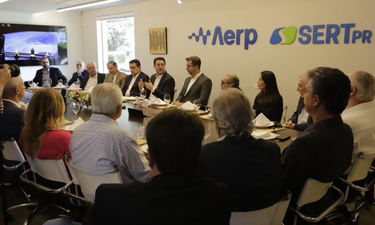 O governador Ratinho Junior fala em uma sala lotada. Ao redor da mesa, membros da Associação das Emissoras de Radiodifusão do Paraná