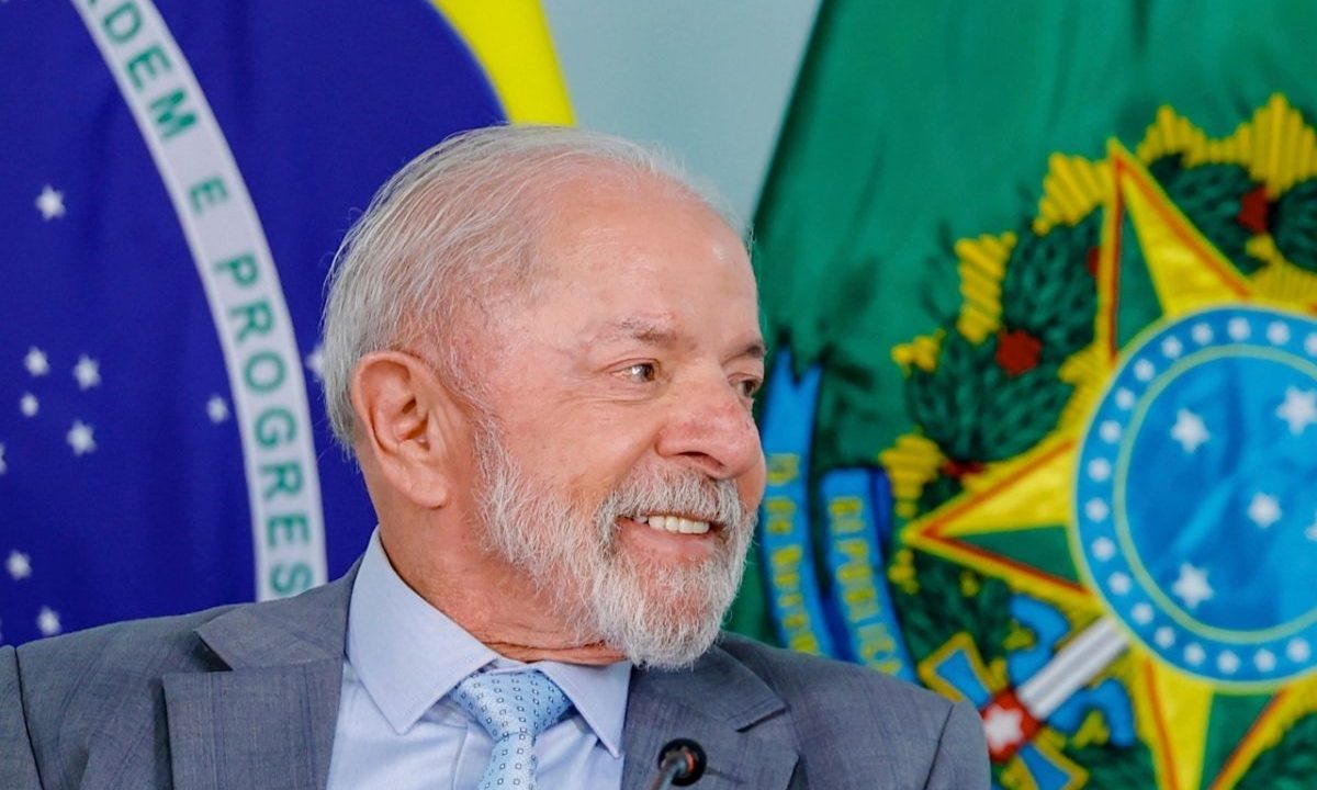 Presidente Lula sorrindo, vestindo terno cinza e gravata azul clara, posicionado em frente às bandeiras do Brasil e das Armas da República.