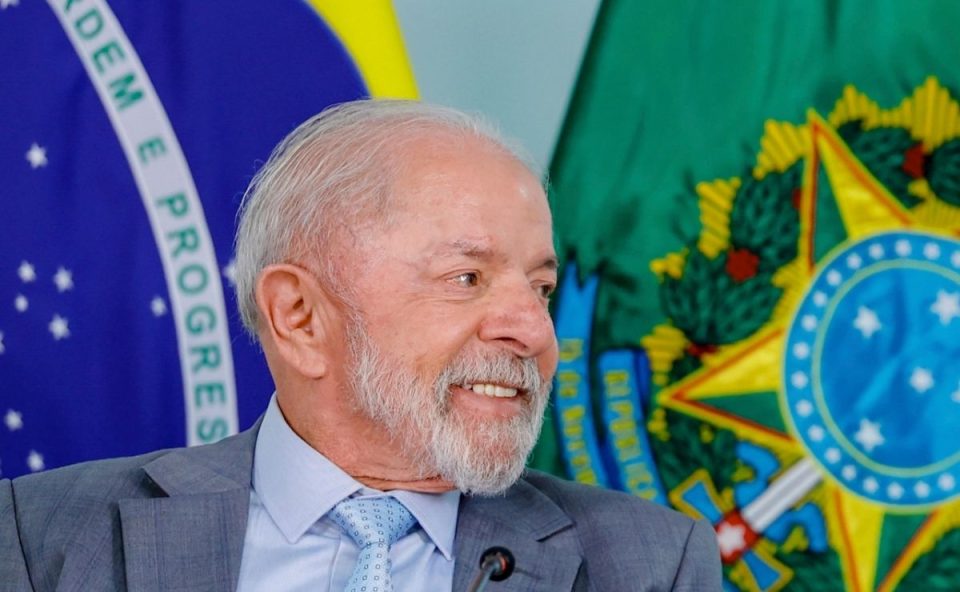 Presidente Lula sorrindo, vestindo terno cinza e gravata azul clara, posicionado em frente às bandeiras do Brasil e das Armas da República.