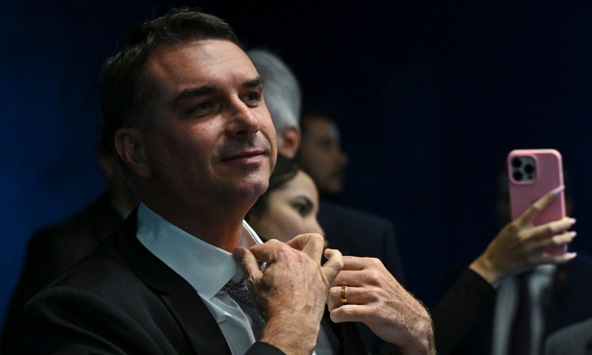 O senador Flávio Bolsonaro, de terno escuro e camisa branca, ajustando o colarinho enquanto olha para cima com um leve sorriso. Ao fundo, uma pessoa segura um celular de cor rosa.