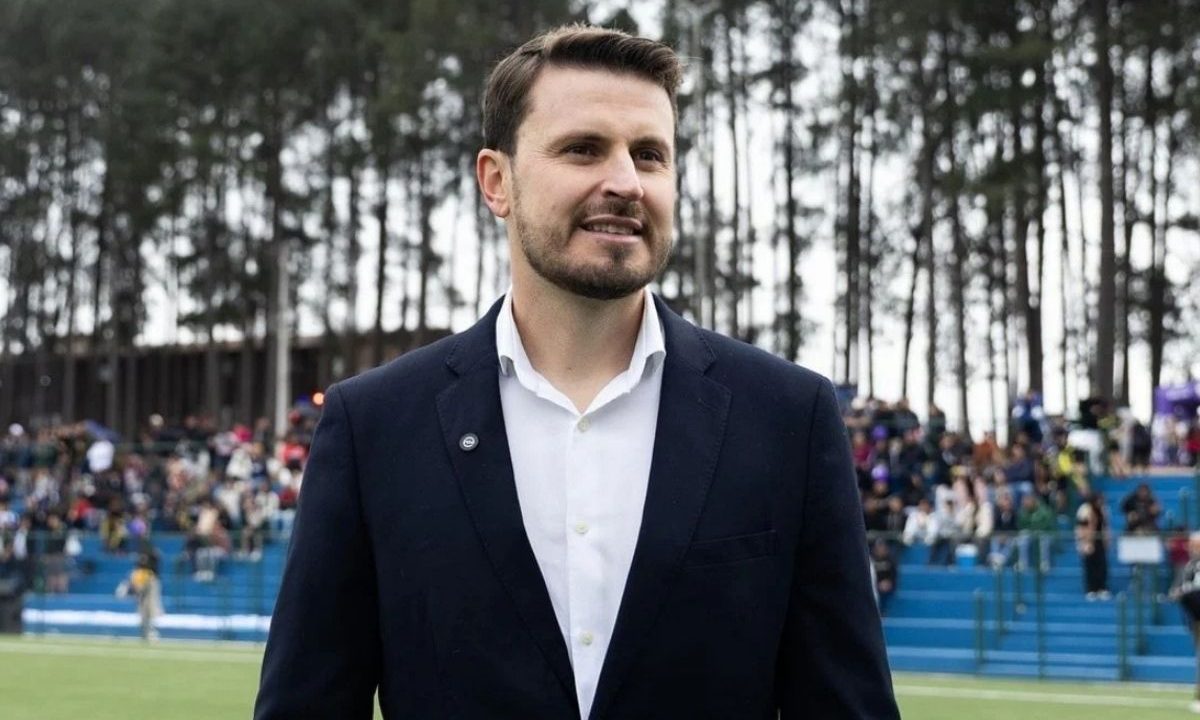 O prefeito Marco Marcondes, de Fazenda Rio Grande, sorri levemente vestindo um blazer azul-marinho e camisa branca. Ele está em um espaço ao ar livre com arquibancadas azuis, público ao fundo e uma linha de pinheiros