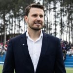 O prefeito Marco Marcondes, de Fazenda Rio Grande, sorri levemente vestindo um blazer azul-marinho e camisa branca. Ele está em um espaço ao ar livre com arquibancadas azuis, público ao fundo e uma linha de pinheiros