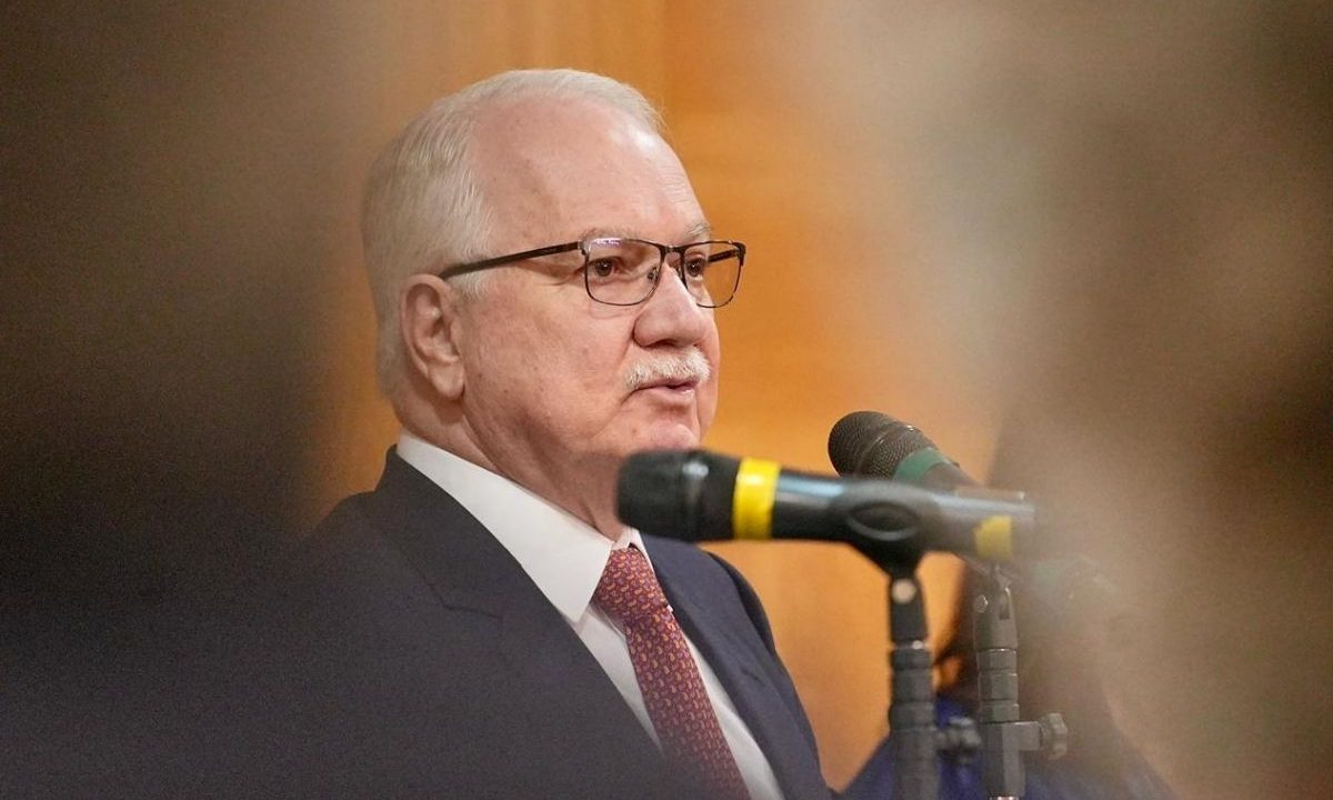 O ministro do STF Edson Fachin, com expressão séria, falando ao microfone durante uma palestra. Ele tem cabelos brancos, usa bigode, óculos de armação escura, terno azul-marinho e gravata vinho com pequenos detalhes. O foco está no ministro, com os microfones em primeiro plano levemente desfocados.