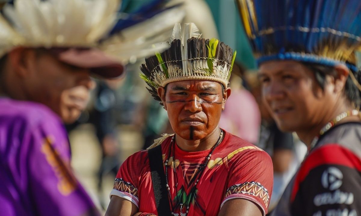 Três homens indígenas da etnia Avá-Guarani. O homem ao centro, em destaque, usa um cocar de penas pretas e brancas com detalhes em verde, pintura facial preta (linhas horizontais no rosto e vertical no queixo) e veste uma camiseta vermelha. Ele olha seriamente para a câmera.