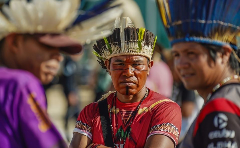 Três homens indígenas da etnia Avá-Guarani. O homem ao centro, em destaque, usa um cocar de penas pretas e brancas com detalhes em verde, pintura facial preta (linhas horizontais no rosto e vertical no queixo) e veste uma camiseta vermelha. Ele olha seriamente para a câmera.