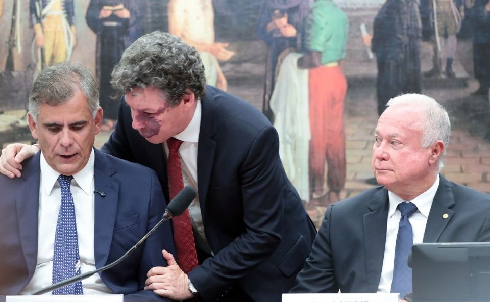 Três homens de terno em uma mesa de comissão. Ao centro, o deputado Reginaldo Lopes está de pé, inclinado, falando ao ouvido do deputado Leur Lomanto Jr, que está sentado à esquerda. À direita, o deputado Paulo Azi observa a conversa.