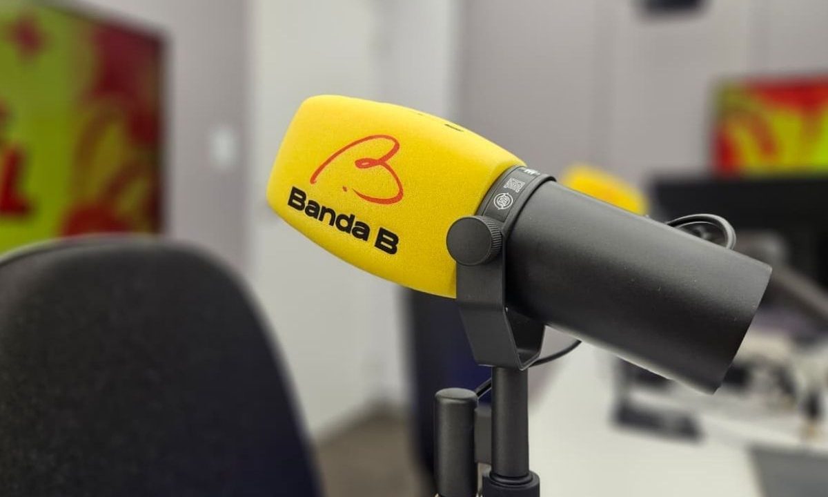 Microfone profissional com espuma amarela exibindo o logotipo da Rádio Banda B em destaque, posicionado dentro de um estúdio com fundo desfocado.