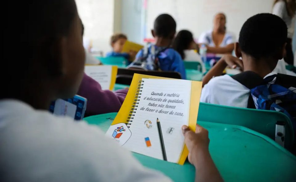 Na imagem, mostra uma criança de costas em uma sala de aula lendo um folheto 