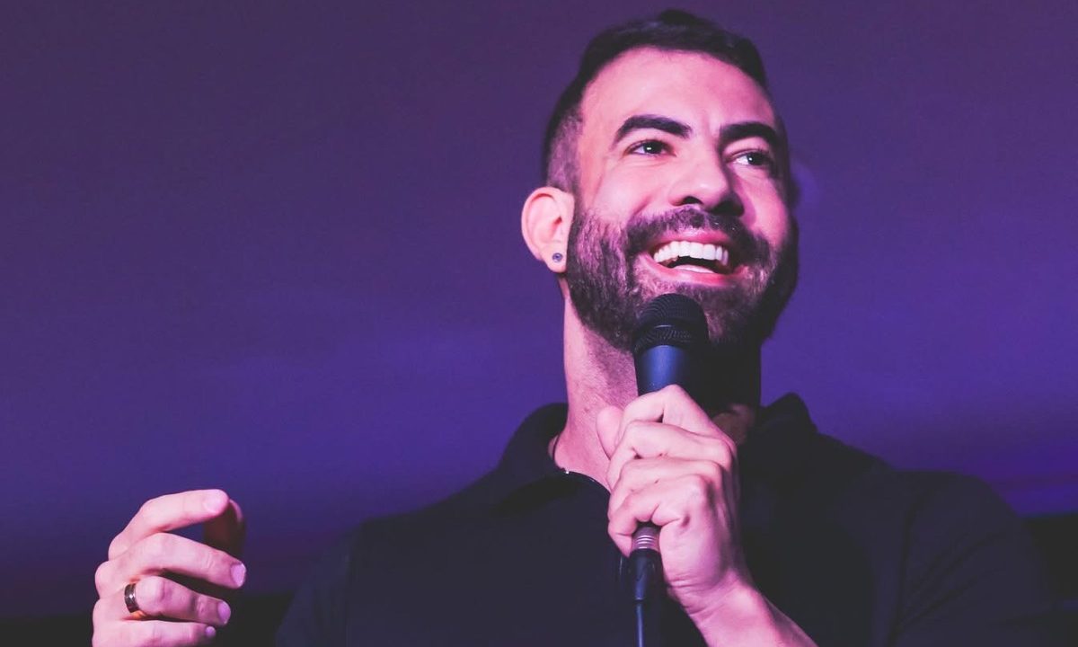 A imagem traz o humorista Raphael Campos que ira trazer o espeta´culo "Fora da Caixinha" a Curitiba. ele está com uma camisa preta, um microfone na mão e sorrindo em cima de um palco