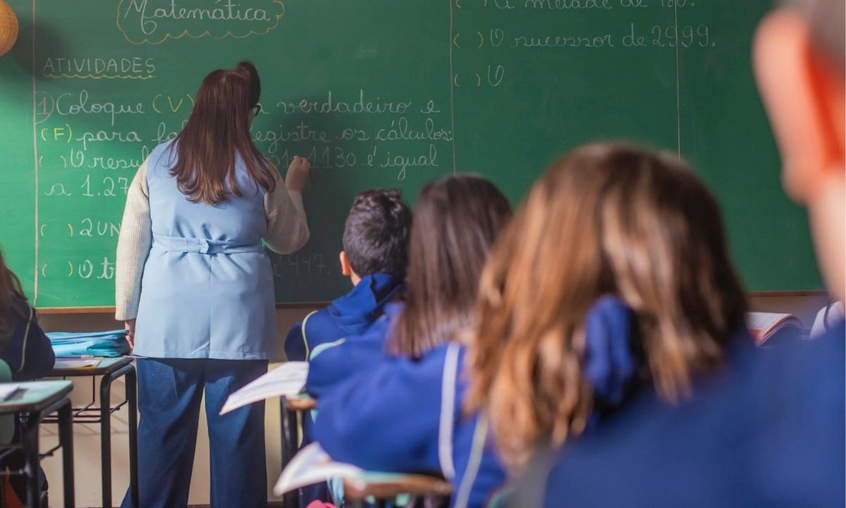 Sala de aula, com uma professora, de costas, escrevendo no quadro negro, e alunos crianças em primeiro plano, também de costas, observando a professora. Professores da rede municipal de Curitiba vão entrar em greve a partir de 8 de abril