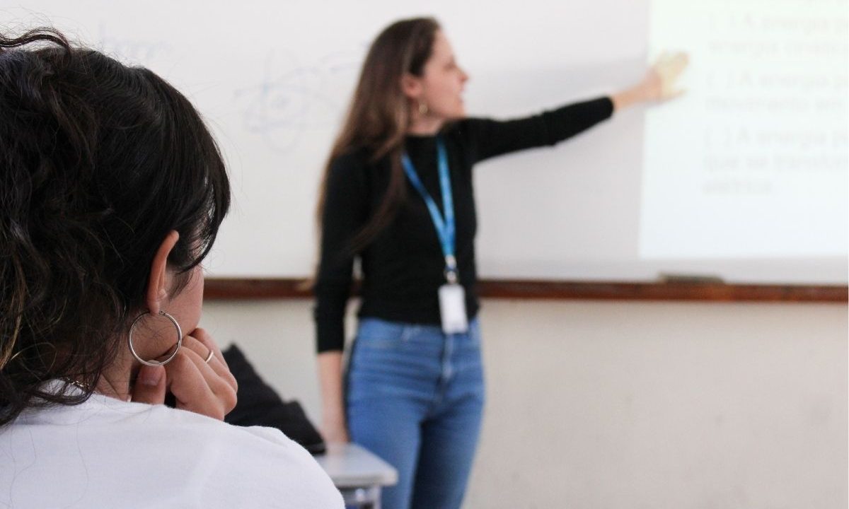 Imagem mostra professora em frente a um quadro. Ela está desfocada. Em primeiro plano está uma aula, de costas. A sala de aula é de um colégio de Santa Catarina. O governador do Estado sancionou uma lei que permite que pais proíbam os filhos de participar de aulas ou atividades pedagógicas sobre orientação ou diversidade sexual e igualdade de gênero