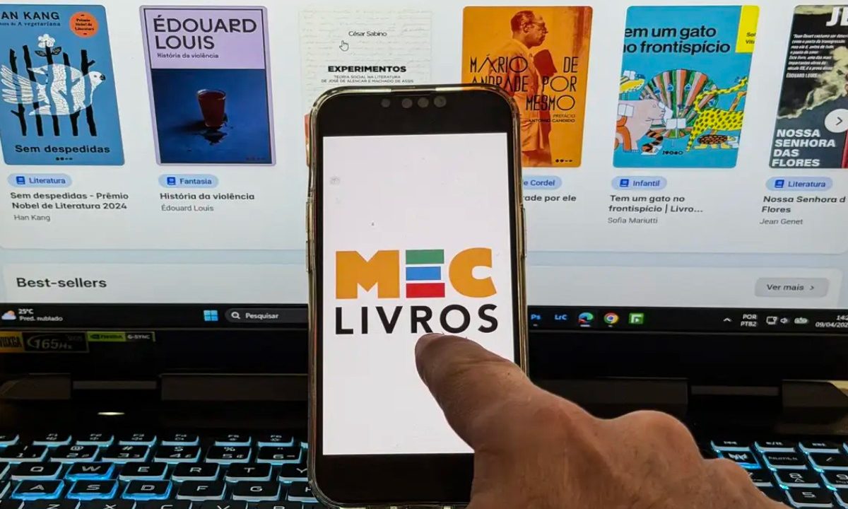 Foto de uma mão mexendo em um celular; na tela tem a logo do aplicativo MEC Livros; ao fundo, um computador logado no aplicativo, apresentando algumas obras do acervo. Imagem ilustrativa para matéria o aplicativo ter ampliado o seu acervo