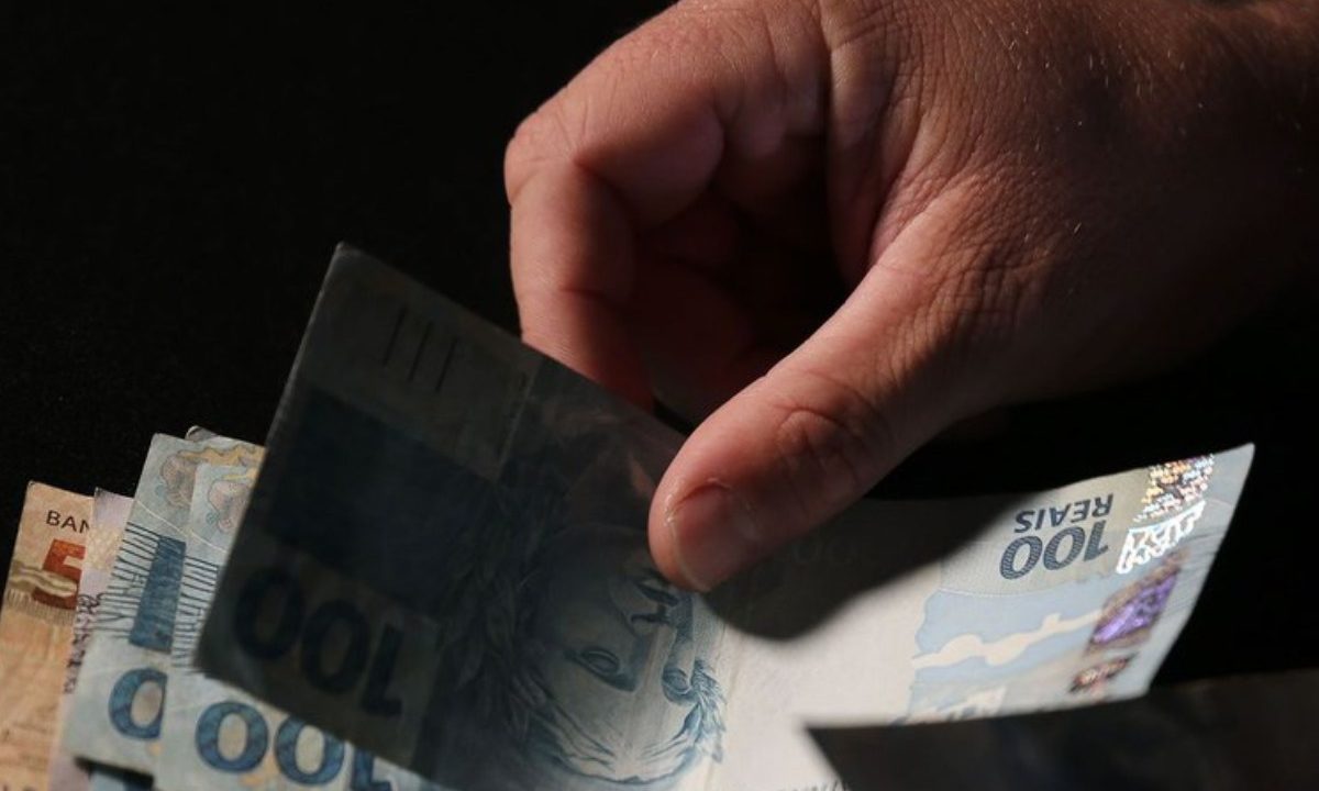 Mão segurando um maço de dinheiro com notas de 100 e 50 reais. Imagem ilustrativa para matéria sobre a primeira parcela do 13º que começa a ser paga pelo INSS nesta semana