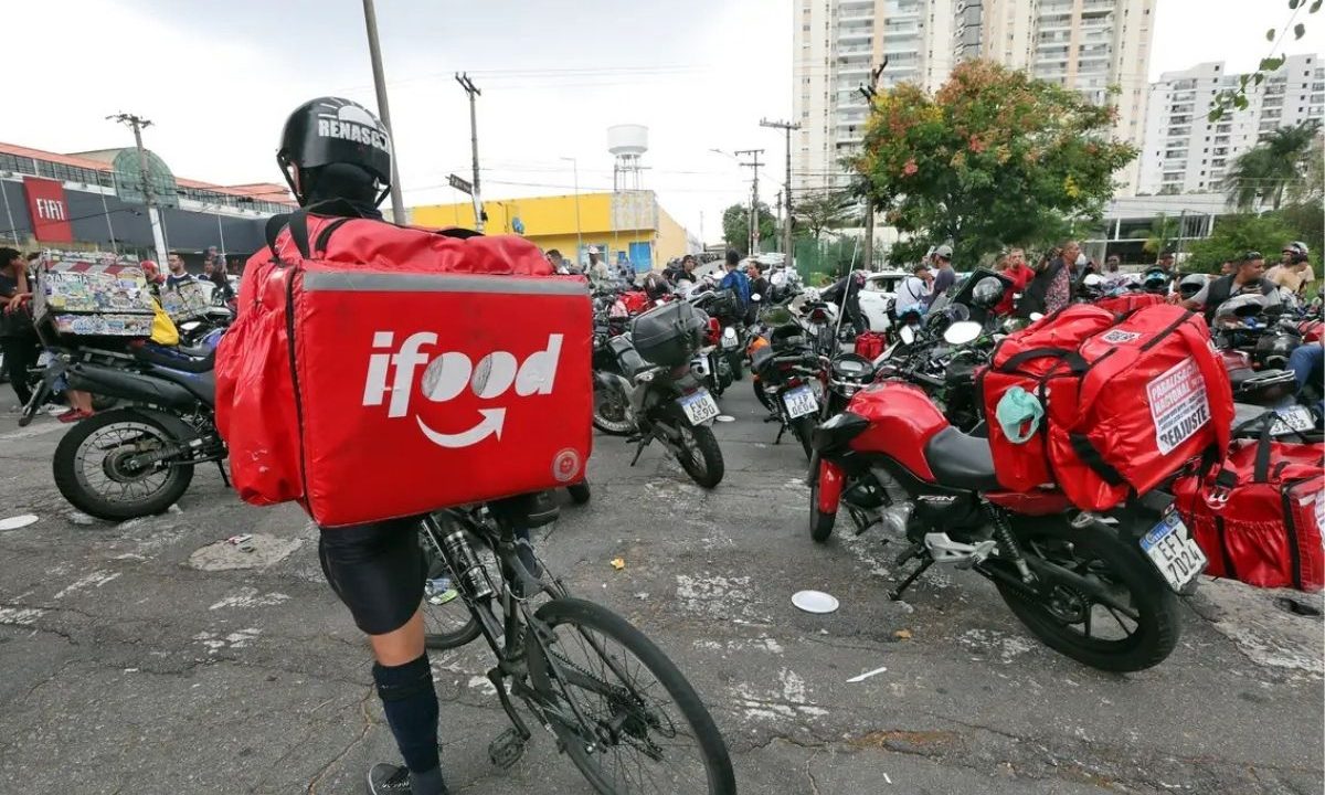 Ciclista entregador do iFood aparece em primeiro plano. Atrás, há entregadores do iFood em motocicletas. A Câmara dos Deputados vai votar o parecer sobre a proposta de regulamentação do trabalho por aplicativo