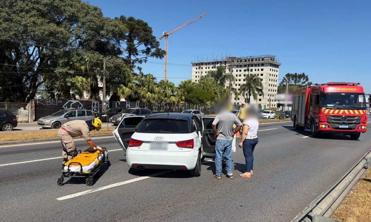 Imagem mostra um acidente na Linha Verde na tarde desta quarta-feira (22), em Curitiba. Um Audi A1 branco está no meio da via. Uma maca espera pela vítima que foi socorrido pelo Siate. Bombeiros trabalham para resgatar a mulher. Duas pessoas estão perto do carro. Um caminhão do Corpo de Bombeiros está atrás na imagem.
