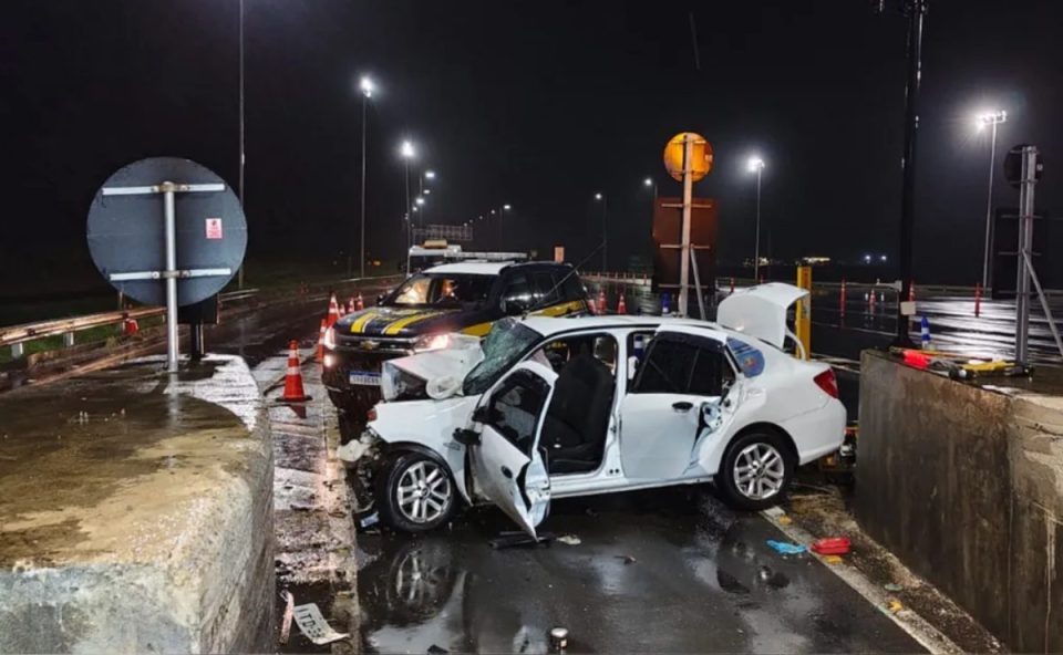 Um acidente provocado por um adolescente, de 17 anos, deixou duas mulheres mortas em Santa Catarina. O condutor do veículo teria perdido o controle e batido de frente com uma mureta em uma praça de pedágio na BR-101. Foto mostra o carro completamente destruído. Atrás, tem uma viatura da PRF parada. O carro do acidente é branco.