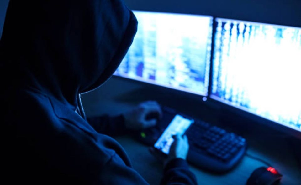 Homem encapuzado em frente a dois monitores e segurando um celular