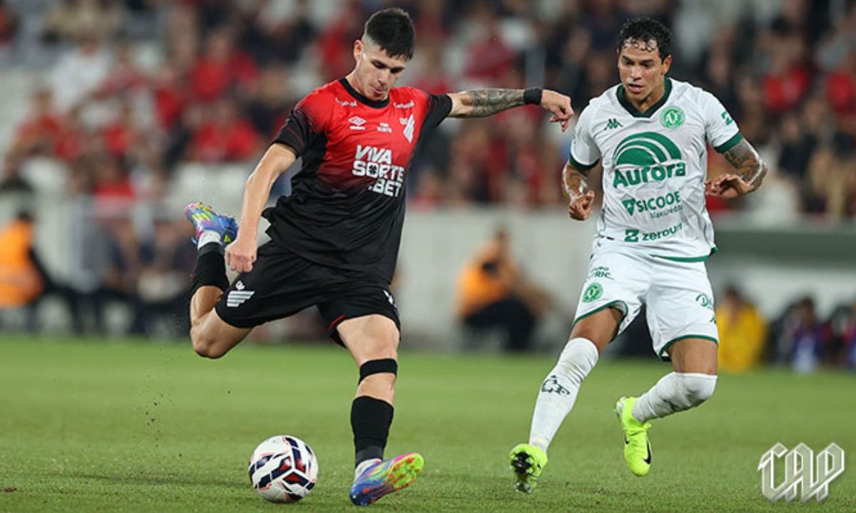 Jogador do Athletico finaliza com a perna direita enquanto é marcado por atleta da Chapecoense durante partida na Arena da Baixada, pelo Campeonato Brasileiro.
