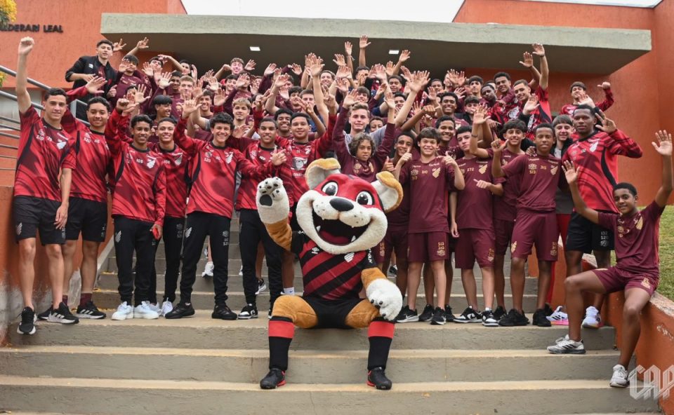 Foto em grupo com dezenas de jogadores das categorias de formação (Sub-15, Sub-17 e Sub-20) posicionados em uma escadaria externa de cor laranja. No centro, à frente de todos, o mascote Fura-Cão está sentado fazendo pose. Todos estão uniformizados.