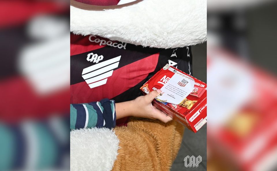 Um close focado em uma criança segurando uma caixa de bombons vermelha. Em cima da caixa, tem um cartão com o escudo do clube escrito
