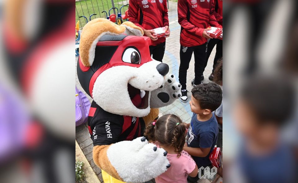 O mascote Fura-Cão, vestindo o uniforme rubro-negro, aparece agachado para falar com uma criança pequena em um pátio escolar. Ao fundo, alguns jogadores da base acompanham a cena.