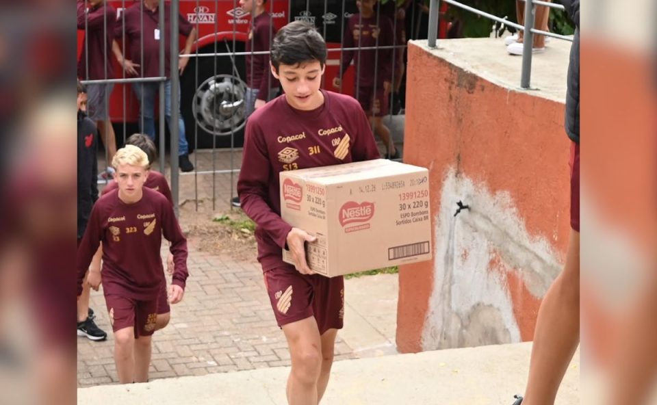 Um dos jogadores da base, de uniforme bordô, desce uma escada carregando uma caixa grande de bombons. Outros atletas vêm logo atrás ajudando no transporte.