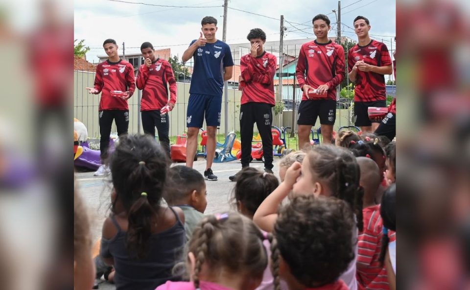 De um ângulo de trás das crianças sentadas, vemos seis jogadores da base em pé conversando com a turma em uma quadra. Um dos atletas sorri e faz um sinal de positivo.