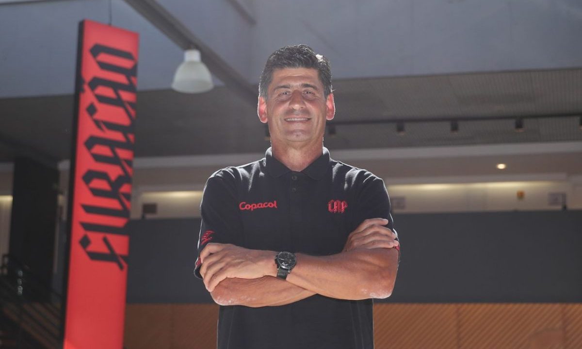 Jorge Raffo, diretor das categorias de base do Athletico, posa com os braços cruzados em ambiente interno do clube, com painel vermelho ao fundo com a palavra “Furacão”