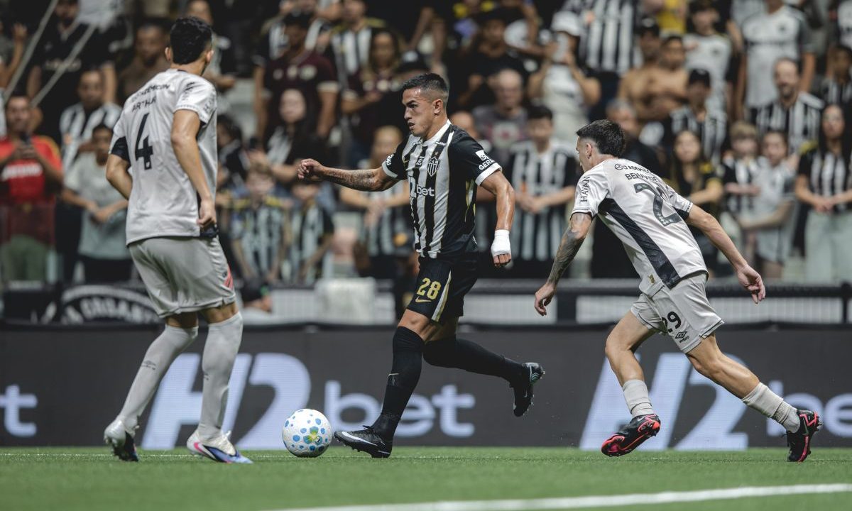 Jogador do Atlético-MG, com uniforme preto e branco, conduz a bola pelo meio de campo enquanto dois atletas do Athletico, com uniforme claro, se aproximam para a marcação. Ao fundo, a torcida acompanha o lance nas arquibancadas da Arena MRV.