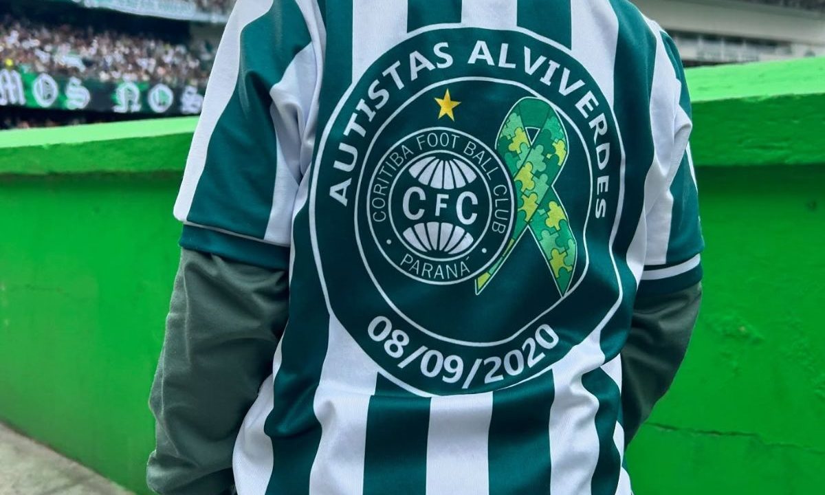 Torcedor do Coritiba aparece de costas vestindo uma camisa verde e branca dentro do estádio Couto Pereira. Na parte de trás do uniforme está estampado o símbolo dos Autistas Alviverdes do Coritiba, com o escudo do clube, um laço com peças de quebra-cabeça — símbolo do autismo — e a data 08/09/2020. Ao fundo, é possível ver parte da arquibancada e o gramado, indicando o ambiente de jogo.