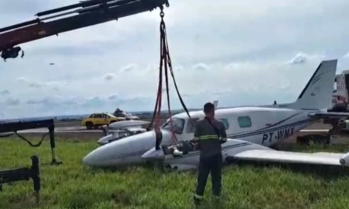 Foto mostra avião de pequeno porte sendo içado por guindaste enquanto trabalhador acompanha o levantamento da aeronave, que perdeu o trem de pouso e saiu da pista com seis pessoas a bordo em Maringá. O avião está sobre a grama.