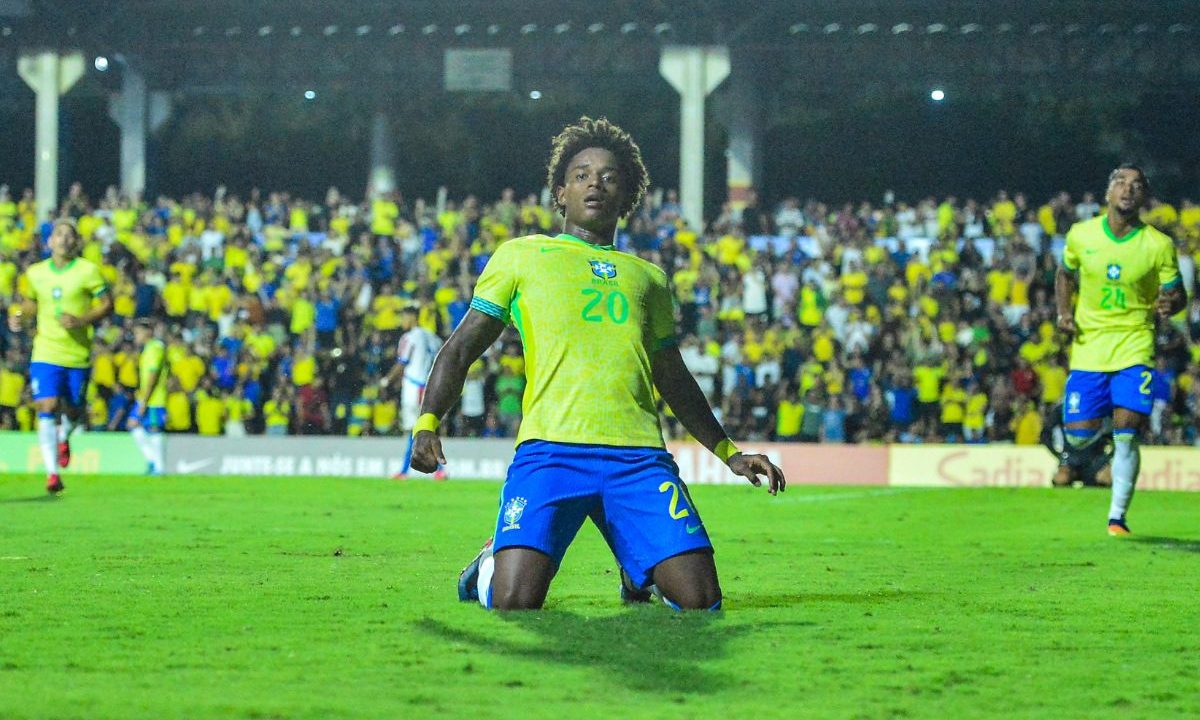 Bruninho, do Athletico, aparece ajoelhado no gramado enquanto comemora um gol pela Seleção Brasileira sub-20. O atacante veste camisa amarela com o número 20, shorts azul e meiões brancos, com os braços levemente abertos e expressão de vibração. Ao fundo, a arquibancada está cheia de torcedores vestindo amarelo, enquanto outros jogadores da seleção aparecem ao redor do campo.