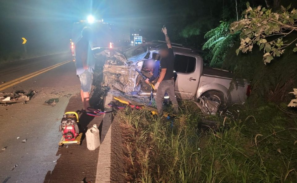 Foto mostra caminhonete destruída após jovem de 24 anos bater de frente com caminhão na BR-153; jovem ficou em estado grave. Foto foi tirada à noite e dois socorristas atendem motorista preso às ferragens, enquanto caminhoneiro observa. Caminhonete está às margens da rodovia