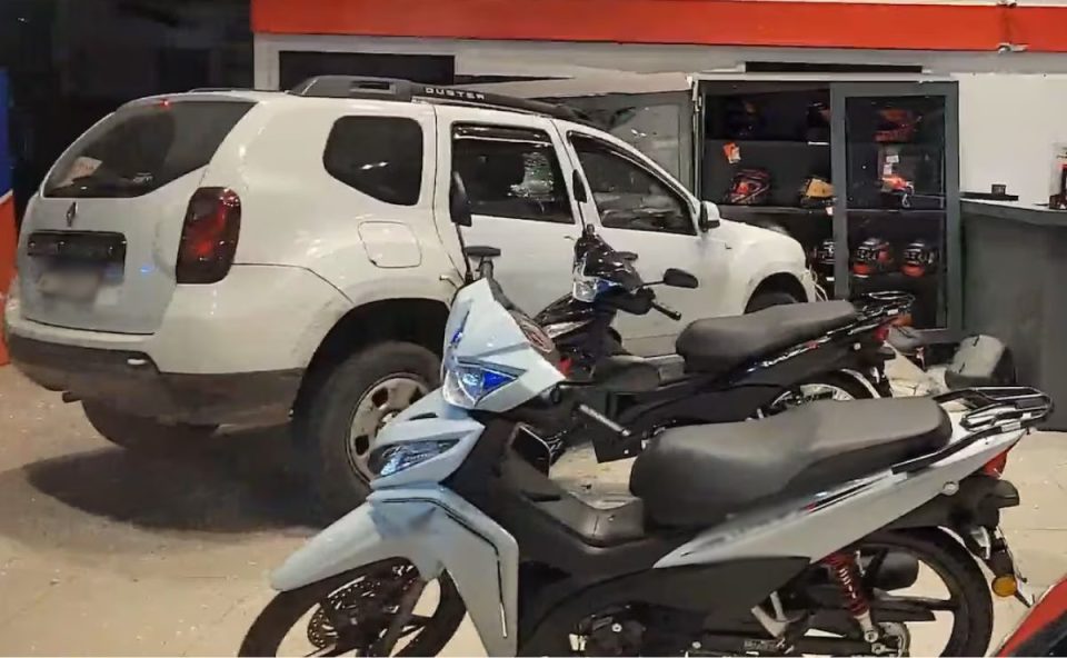 Imagem mostra o carro que bateu na moto e invadiu concessionária dentro da loja. O veículo envolvido no acidente é uma Renault Duster branca. Ela está posicionada dentro da loja, ao lado de algumas motos e perto de um móvel totalmente quebrado.