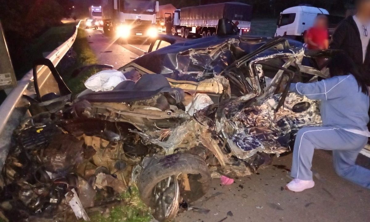 Foto tirada durante a noite mostra carro de casal e criança da mesma família destruído após eles se envolverem em colisão frontal com carreta na BR-116, em Rio Negro, na Região Metropolitana de Curitiba. Família morreu no acidente. Trânsito flui com lentidão na foto, e testemunhas se aproximam do carro