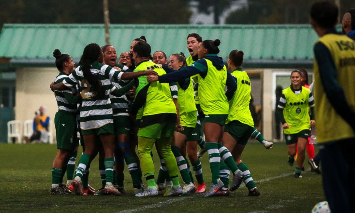 Atletas do Coritiba feminino, vestindo uniforme verde e branco, se abraçam e comemoram em grupo dentro de campo após um gol, durante partida disputada sob clima chuvoso.