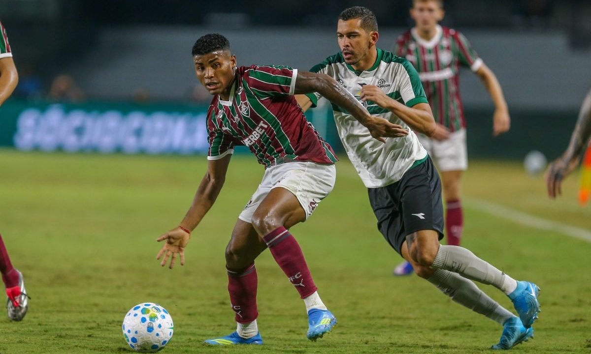 Dois jogadores disputam a bola durante o jogo entre Coritiba e Fluminense no Couto Pereira. O atleta do Fluminense, com camisa tricolor grená, verde e branca, protege a bola enquanto é pressionado por um jogador do Coritiba, que veste uniforme branco e verde. A jogada acontece em velocidade, com ambos inclinados para frente e focados na posse.
