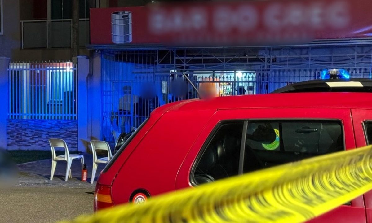 Foto tirada durante à noite mostra corpo de empresário executado a tiros em frente a bar. Ele está caído sobre uma calçada, ao lado de uma mesa branca de plástico. Policial militar e guarda municipal conversam enquanto observam o corpo. Um carro vermelho ocupa parte da foto, em primeiro plano.