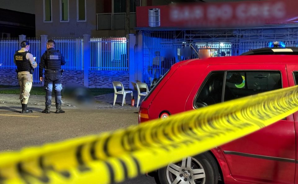 Foto tirada durante à noite mostra corpo de empresário executado a tiros em frente a bar. Ele está caído sobre uma calçada, ao lado de uma mesa branca de plástico. Policial militar e guarda municipal conversam enquanto observam o corpo. Um carro vermelho ocupa parte da foto, em primeiro plano.