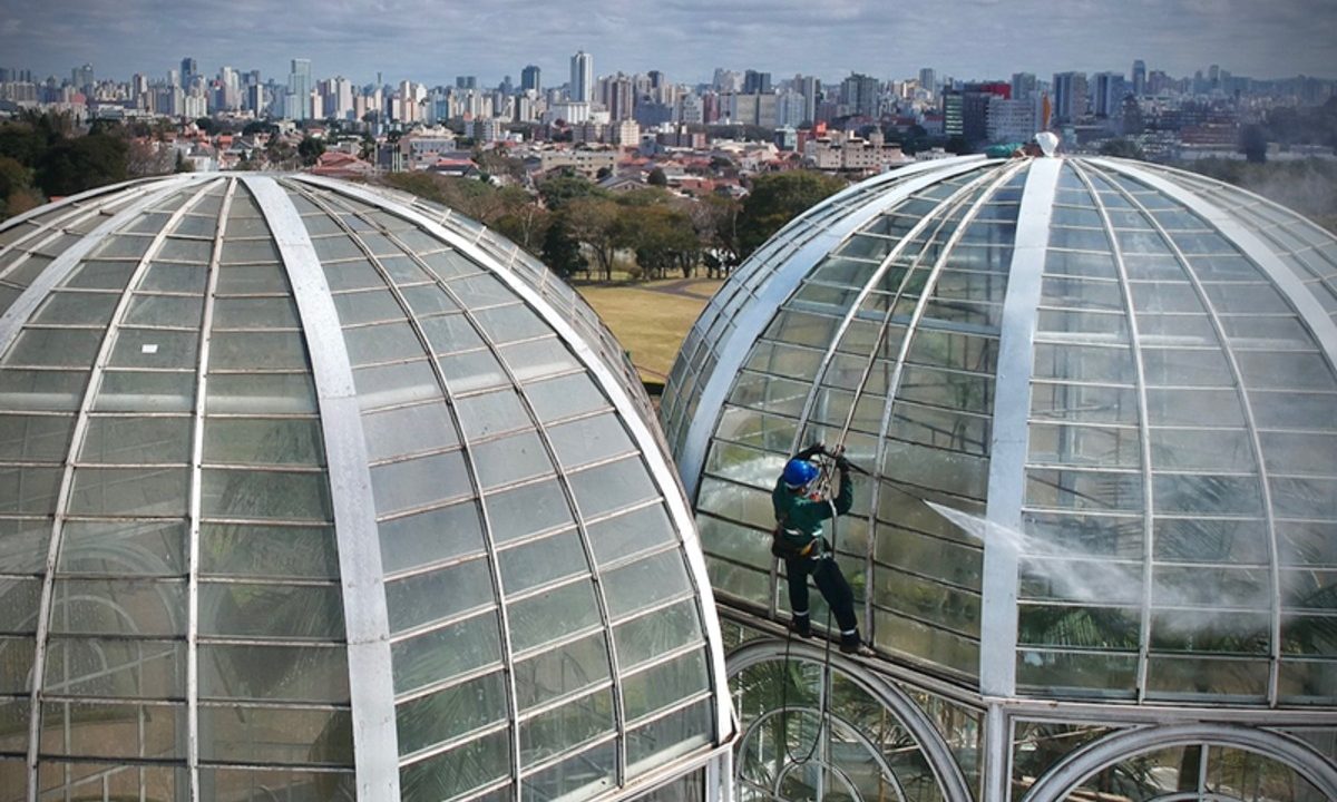 Foto mostra trabalhador no alto da estrutura do Jardim Botânico de Curitiba fazendo limpeza de vidros, que volta a ser fechada para a realização da manutenção
