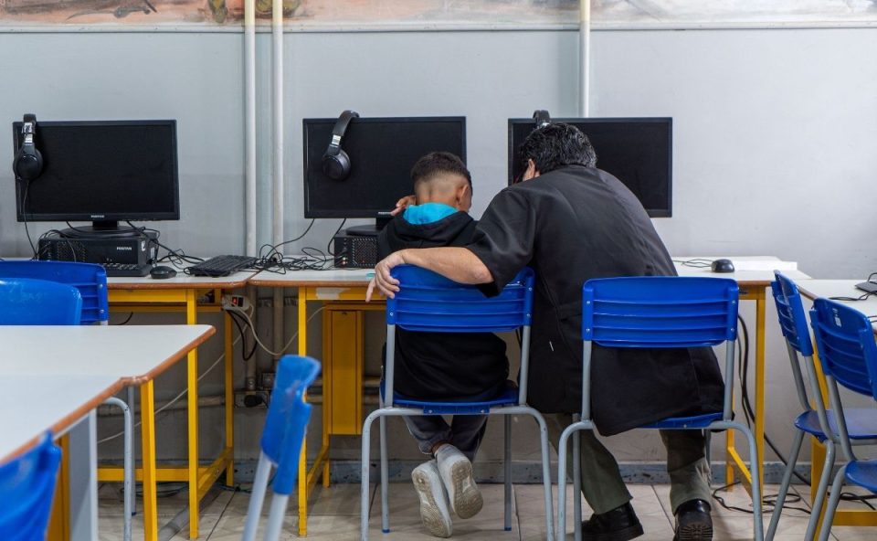 Um adulto e um jovem estudante sentados lado a lado em um laboratório de informática de uma escola estadual. O adulto, de costas para a câmera, está com o braço apoiado na cadeira do estudante, em um gesto de acompanhamento e apoio. Há monitores de computador e cadeiras azuis no ambiente.