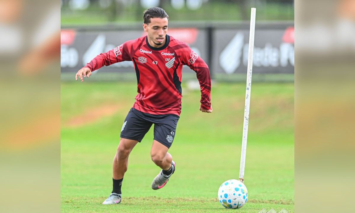 Felipe Chiqueti do Athletico aparece em campo durante um treinamento, vestindo uniforme vermelho e preto do clube. O atacante conduz a bola em movimento, com o corpo inclinado para frente e olhar focado, enquanto realiza um exercício com estaca de treino no gramado do CT do Caju.
