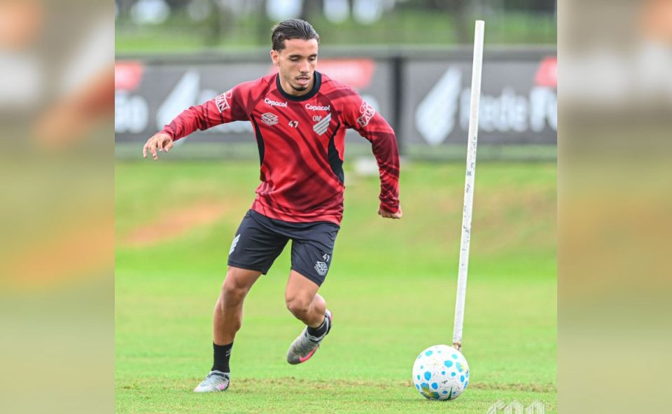 Felipe Chiqueti do Athletico aparece em campo durante um treinamento, vestindo uniforme vermelho e preto do clube. O atacante conduz a bola em movimento, com o corpo inclinado para frente e olhar focado, enquanto realiza um exercício com estaca de treino no gramado do CT do Caju.