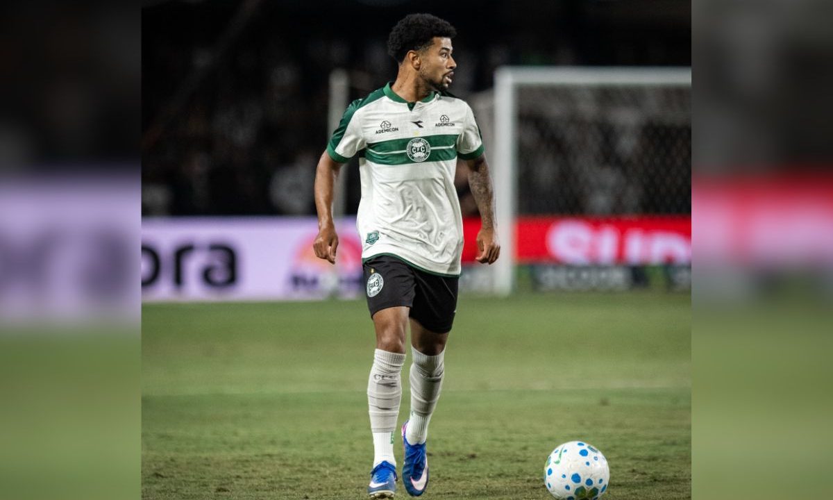 Lucas Ronier, jogador do Coritiba, conduz a bola durante partida no Couto Pereira, usando uniforme branco e verde, em lance de jogo.