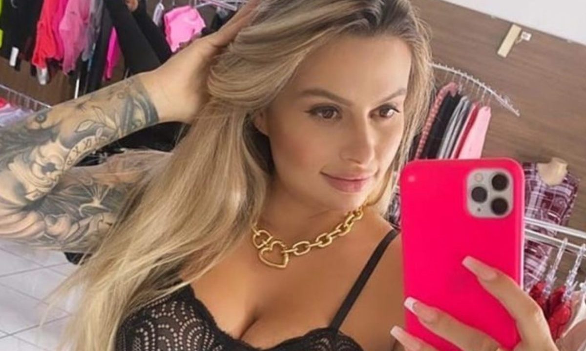 Trafigata, em prisão domiciliar, conta sobre própria história. Ela é loira, está com a mão no cabelo e com top preto. A foto foi tirada na frente do espelho. Ela segura o celular com capinha rosa.