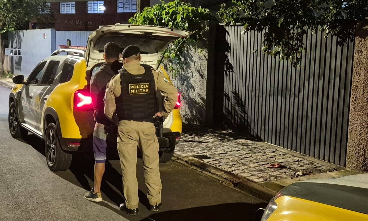 Imagem mostra homem condenado por matar cunhado que defendeu irmã em Campo Largo sendo algemado por um policial militar próximo a uma viatura. Ele aparece de costas, usando moletom e bermuda roxa, após ser responsabilizado pela morte do cunhado, que foi atingido ao tentar proteger a irmã durante um ataque a facadas.