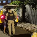 Imagem mostra homem condenado por matar cunhado que defendeu irmã em Campo Largo sendo algemado por um policial militar próximo a uma viatura. Ele aparece de costas, usando moletom e bermuda roxa, após ser responsabilizado pela morte do cunhado, que foi atingido ao tentar proteger a irmã durante um ataque a facadas.