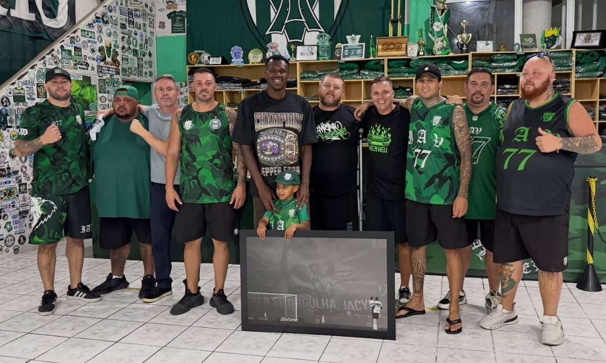Jacy posa ao lado de integrantes da torcida organizada Império Alviverde dentro da sede, segurando um quadro com imagem sua em campo com o filho no colo e um bandeirão ao fundo em homenagem à sua mãe