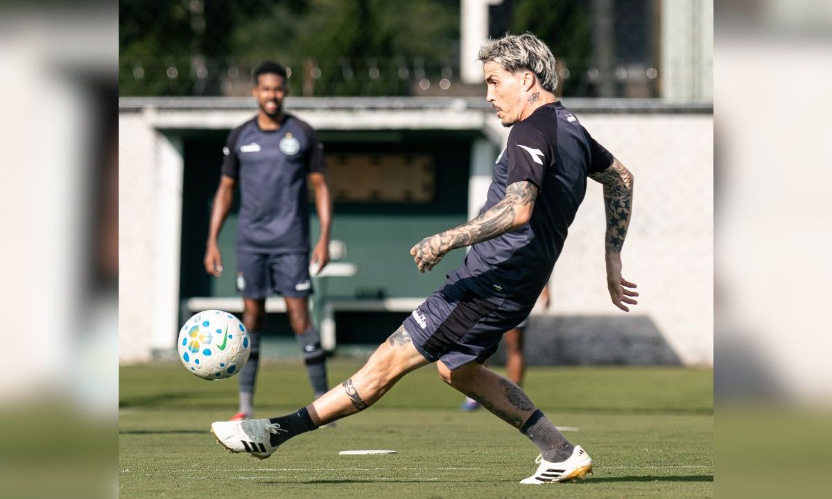 Josué do Coritiba aparece em campo durante um treinamento, vestindo uniforme escuro do clube. O meia está no momento do passe, com a perna estendida tocando a bola, enquanto outro jogador observa ao fundo. O gramado está bem cuidado e o treino acontece em ambiente aberto, com estrutura do centro de treinamento ao fundo.